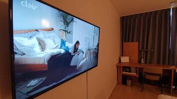 A large flat-screen TV mounted on a wall displaying a room with a bed, a plant, and a desk with a chair.