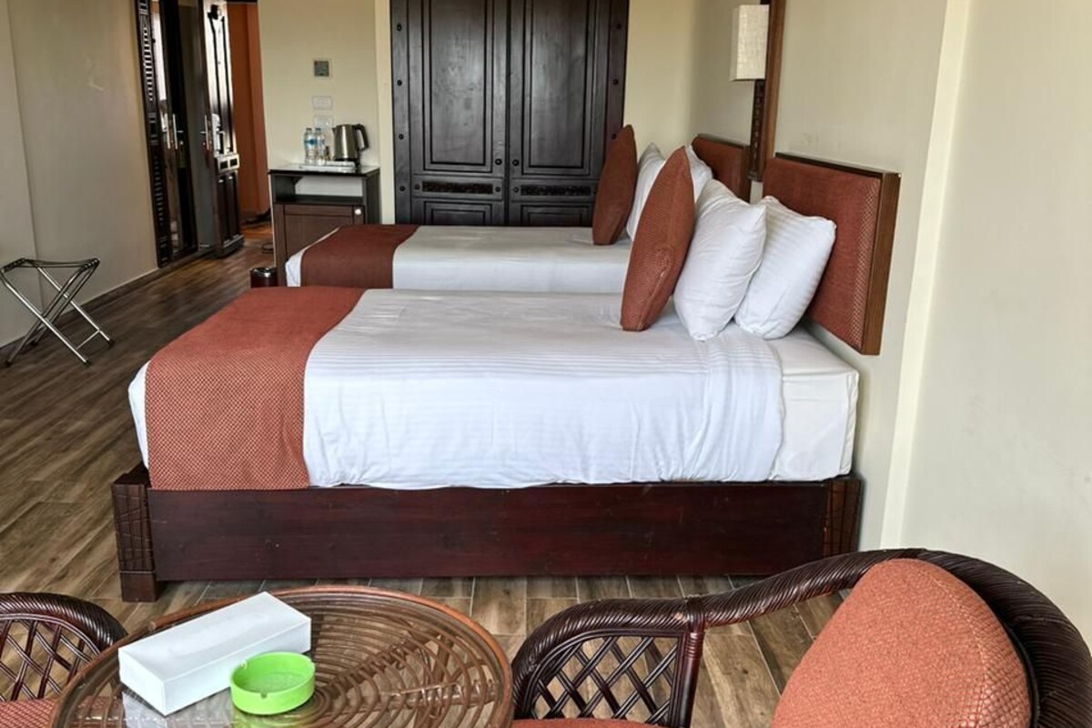 Habitación de hotel con dos camas, una mesita con un tazón verde, sillas de mimbre y un armario de madera.