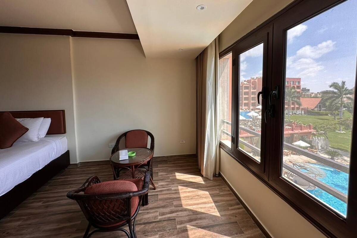 A hotel room with a bed, a dining table with chairs, and a large window with a view of a pool and buildings.