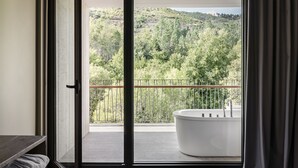 A modern bathroom with a large bathtub, a balcony with a metal railing, and a view of a forested hillside. - Aqua Village Health Resort & Spa (Oliveira do Hospital)
