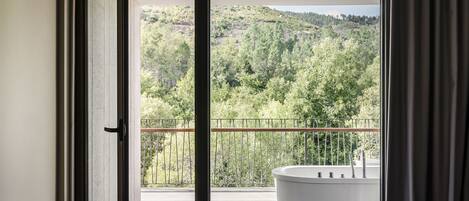 A modern bathroom with a large bathtub, a balcony with a metal railing, and a view of a forested hillside.