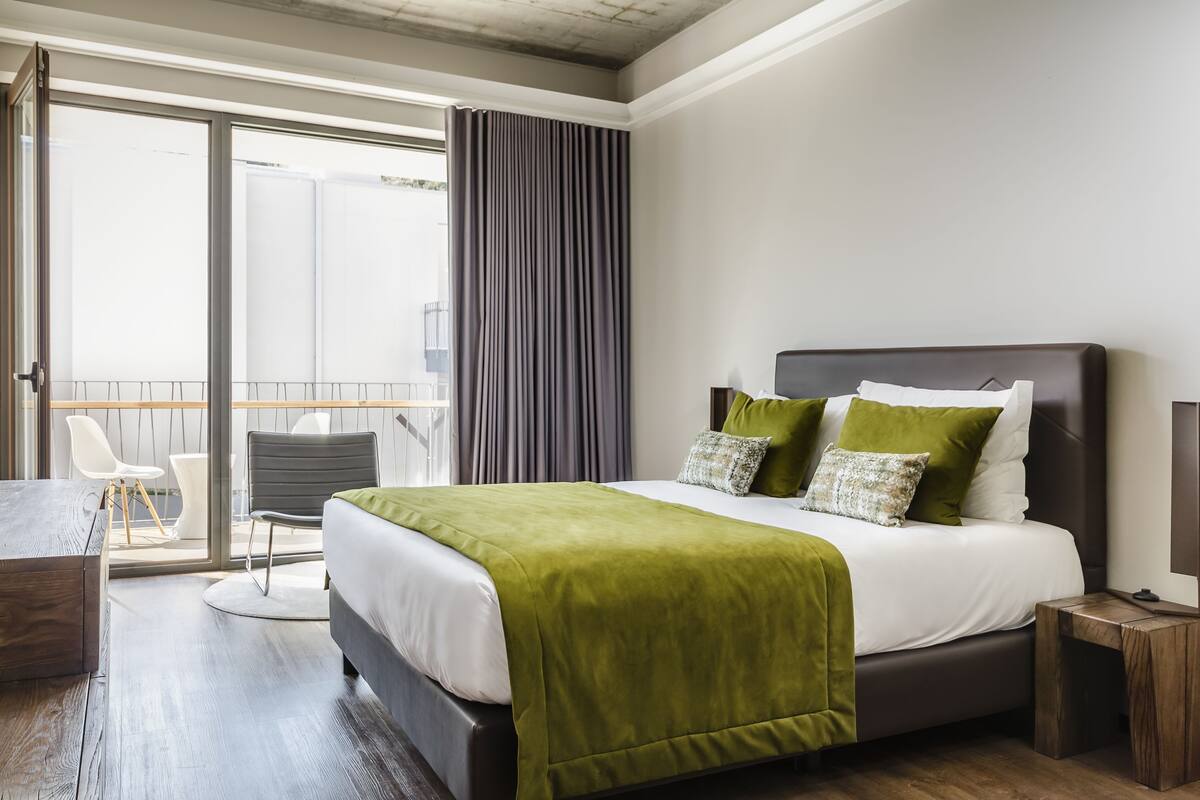 Een moderne hotelkamer met een groot bed, een balkon met stoelen en uitzicht op de stad.