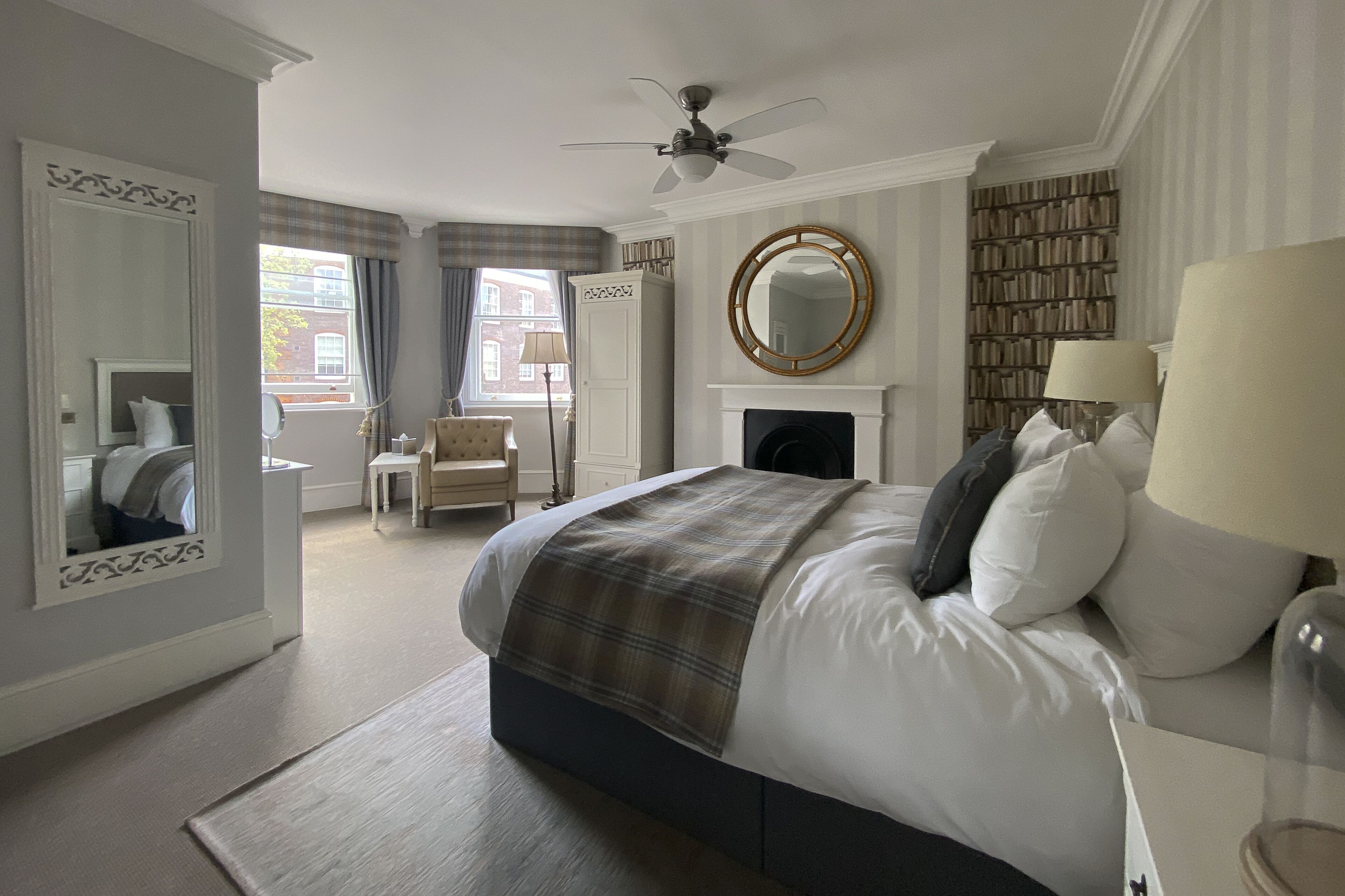 A bedroom with a bed, a fireplace, a ceiling fan, a mirror, a bookshelf, and a window with curtains.