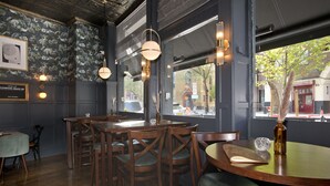 A restaurant interior with wooden tables and chairs, a large window with a view of the street, and decorative wall sconces. - The Clerk & Well Pub & Rooms (London)