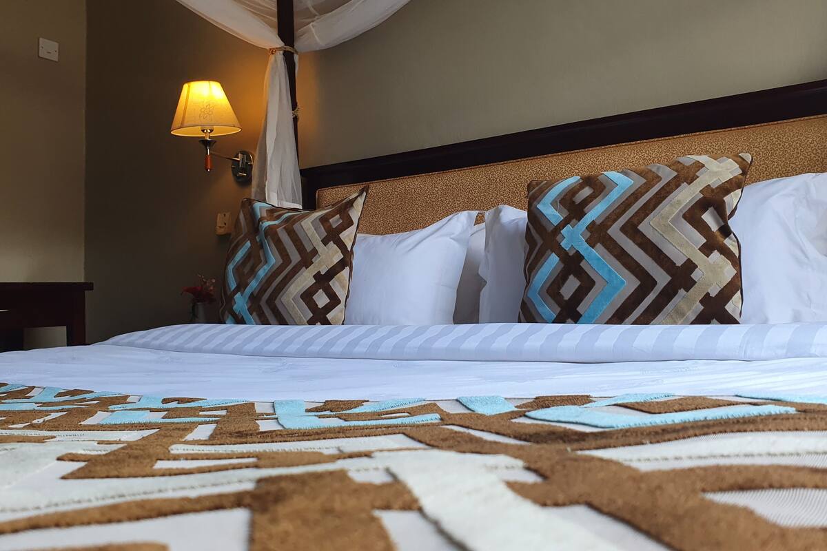 Una habitación de hotel con cama, colcha estampada, almohadas, una mesita de noche, una lámpara y un aplique en la pared.