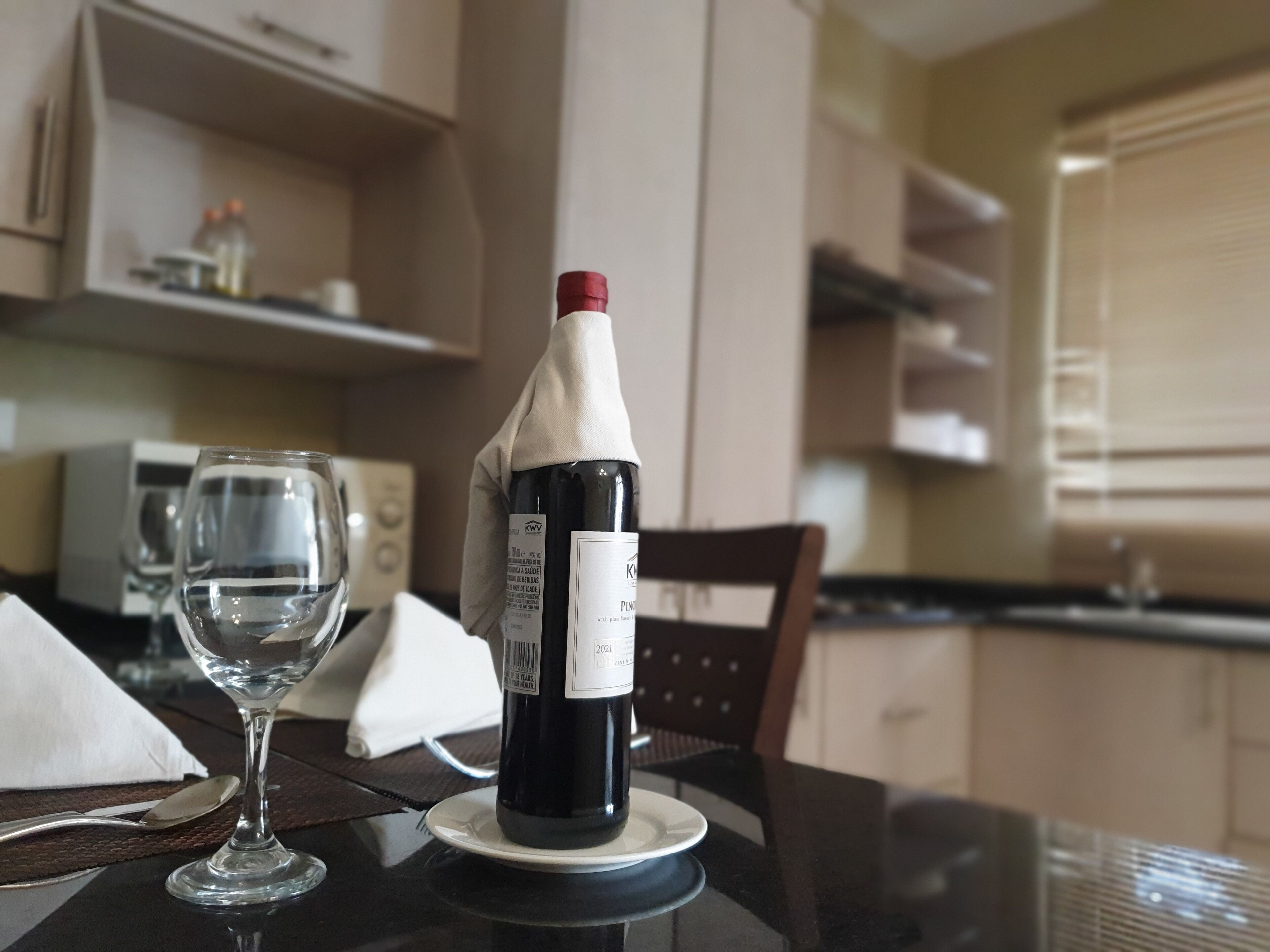 A bottle of wine on a coaster with a glass and a napkin on a table in a kitchen setting.
