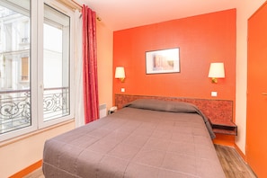 Ett hotellrum med en stor säng, två sänglampor, ett fönster med utsikt över byggnader och en orange accentvägg.