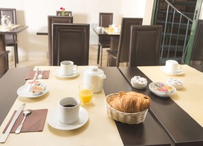 A breakfast setup with a croissant basket, coffee cups, a glass of orange juice, and plates with pastries on a hotel dining table. - Hôtel Pierre Nicole (Paris)