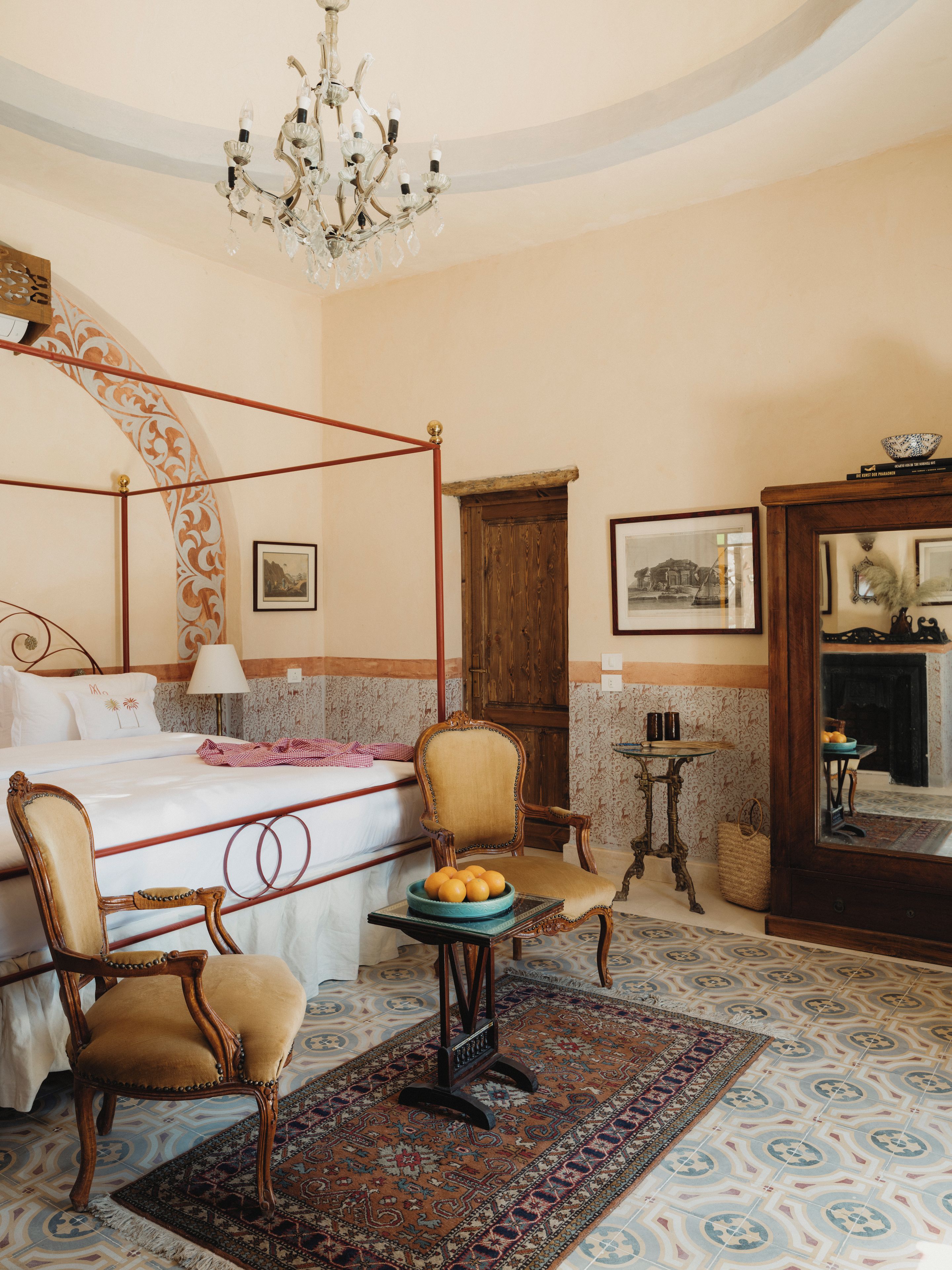 A bedroom with a four-poster bed, a chandelier, a wooden door, and a patterned rug.