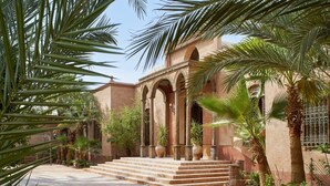 A courtyard with a building featuring arches and columns, surrounded by palm trees and potted plants. - Al Moudira Hotel (Luxor)
