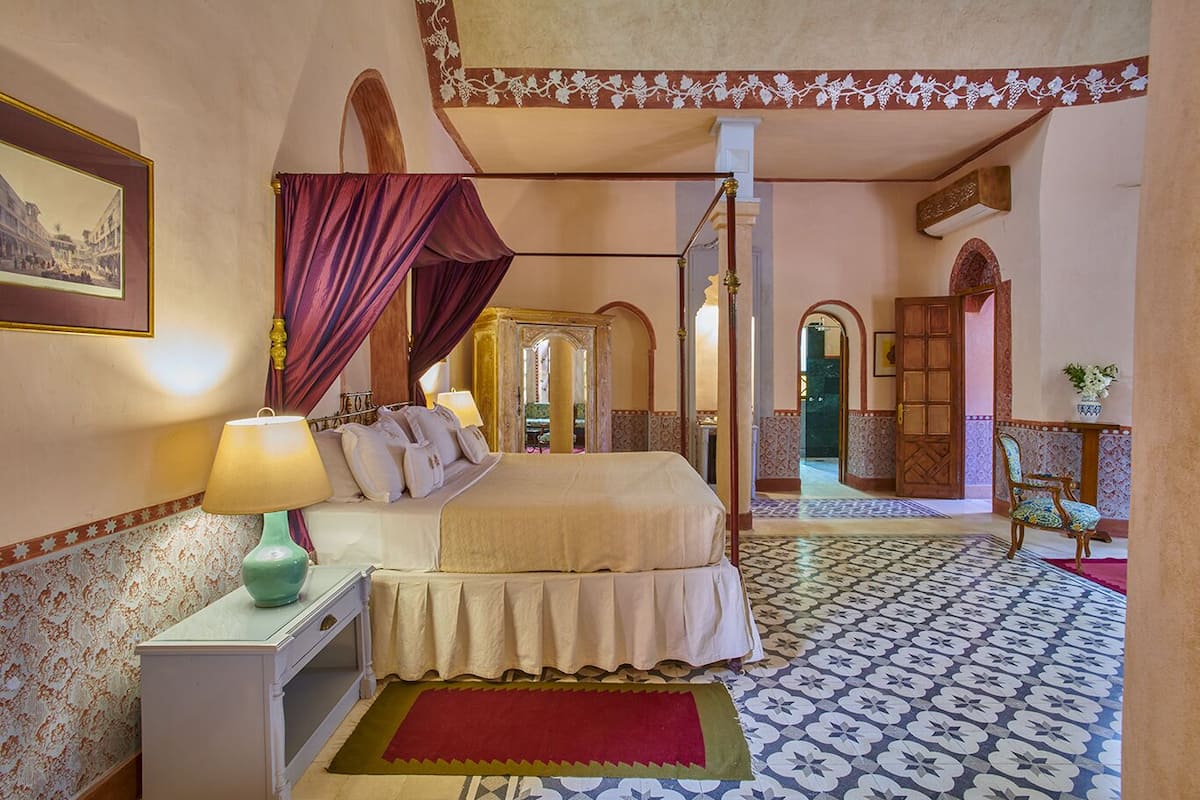 A four-poster bed with a canopy, a nightstand with a lamp, a red rug, and a patterned floor.