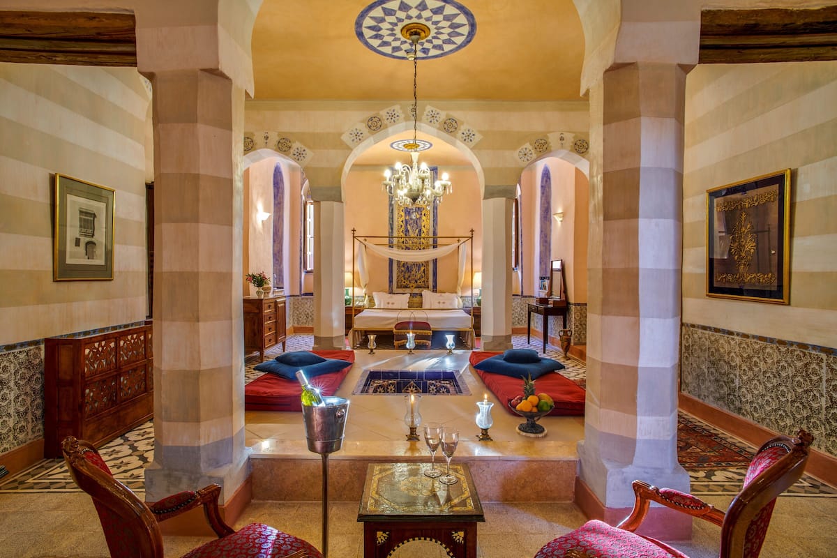 A luxurious hotel lobby with a central seating area, a chandelier, and a view of the bedroom.