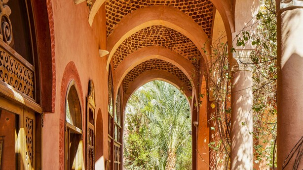 An arched corridor with a view of a garden, wooden furniture, and a tiled floor. - Al Moudira Hotel (Luxor)