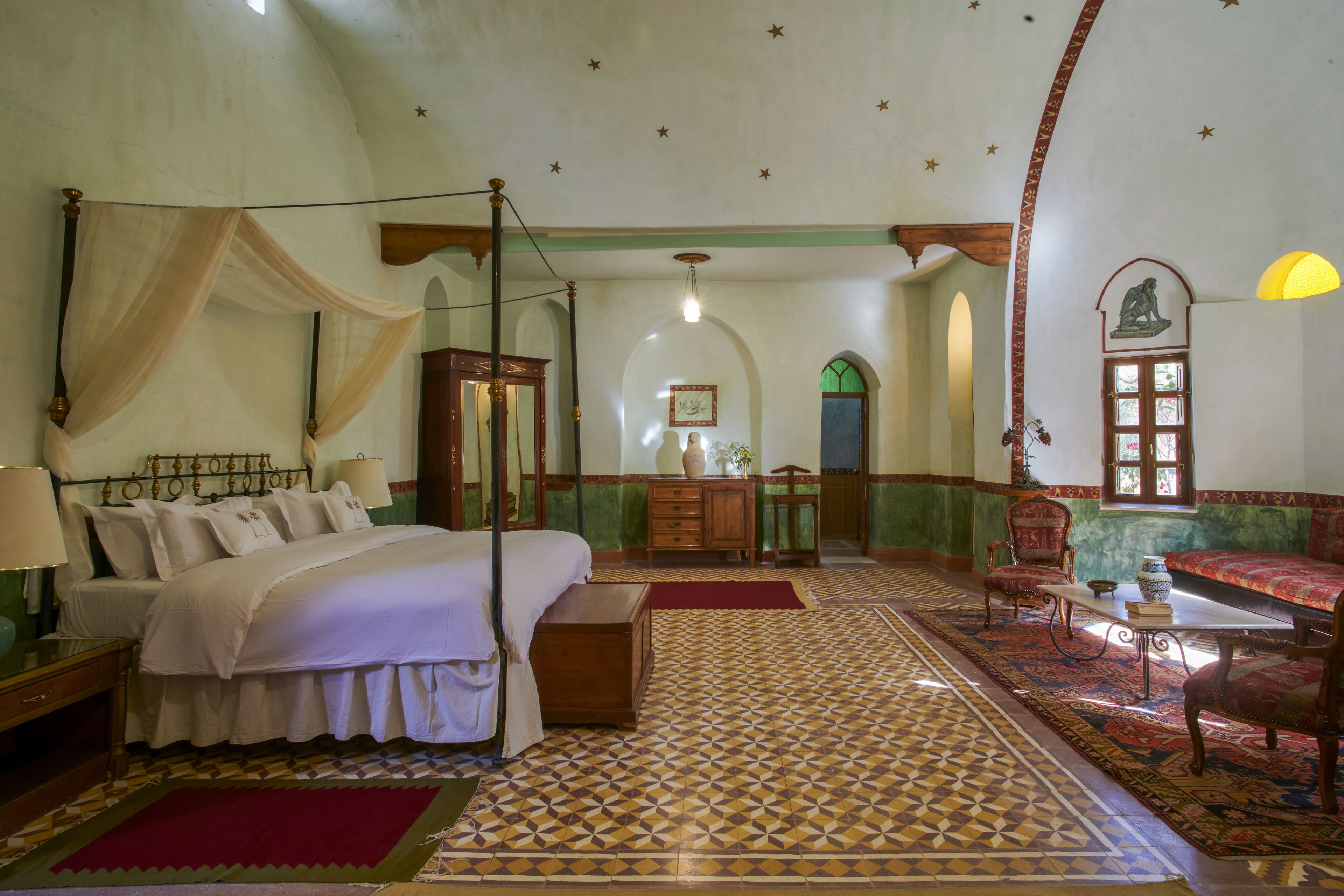 A bedroom with a four-poster bed, a wooden dresser, a red rug, and a patterned floor.