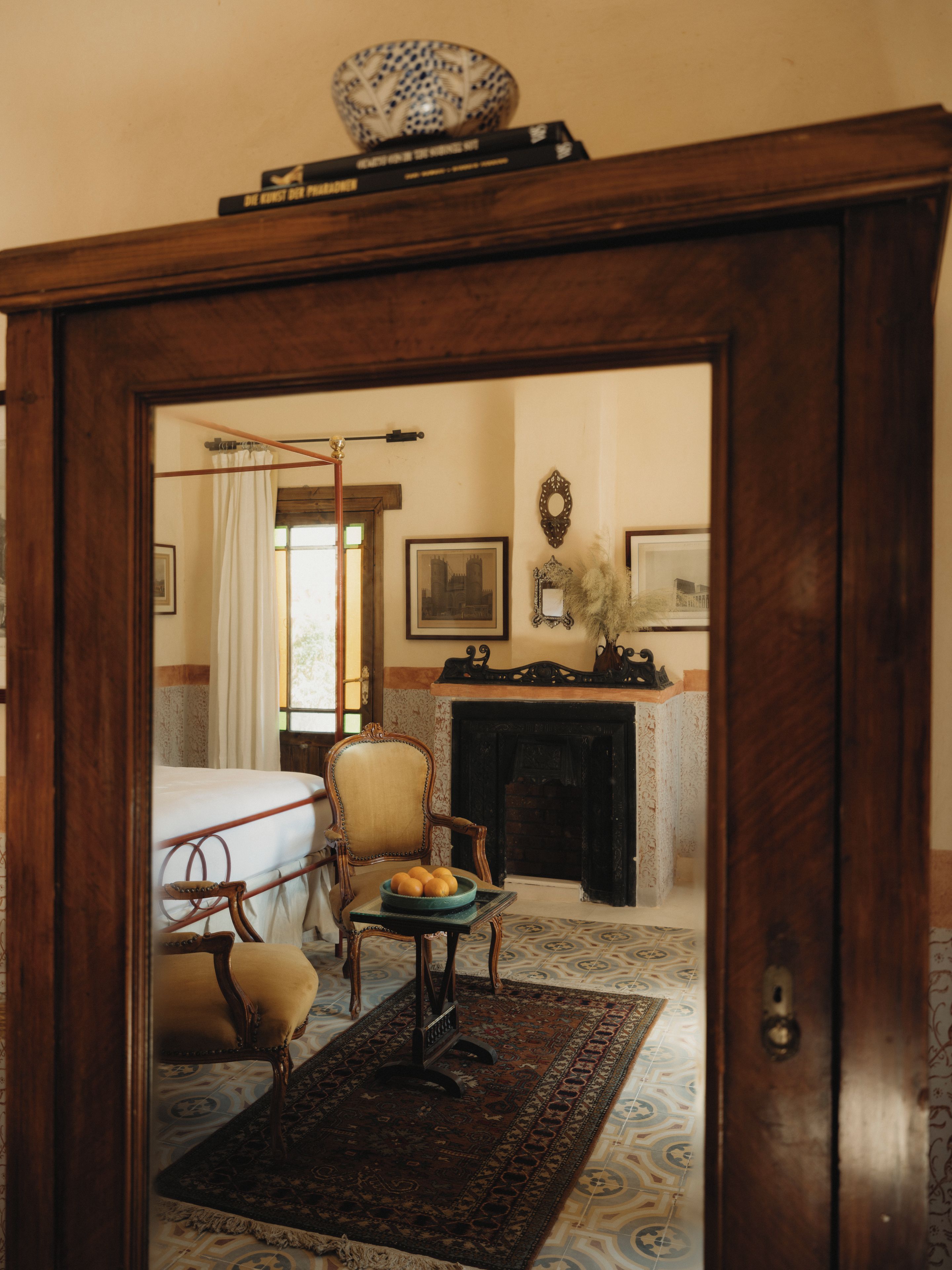 A room with a bed, a chair, a table with fruit, a fireplace, and a mirror.