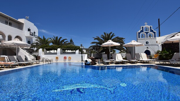 Una piscina azul cristalina con sillas de descanso y sombrillas, rodeada de edificios blancos con una torre de campanas.