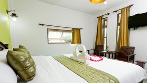 Una habitación de hotel con una cama bien hecha, un plaid verde, una decoración con forma de cisne blanco, una silla de madera y un televisor montado en la pared.