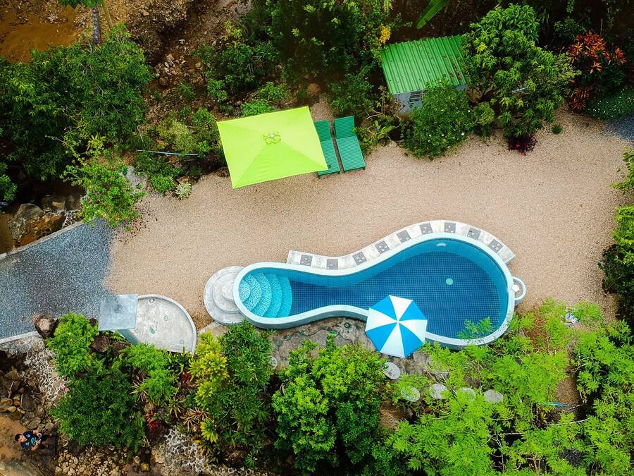 An aerial view of a swimming pool surrounded by lush greenery, with a small bridge leading to the pool area.