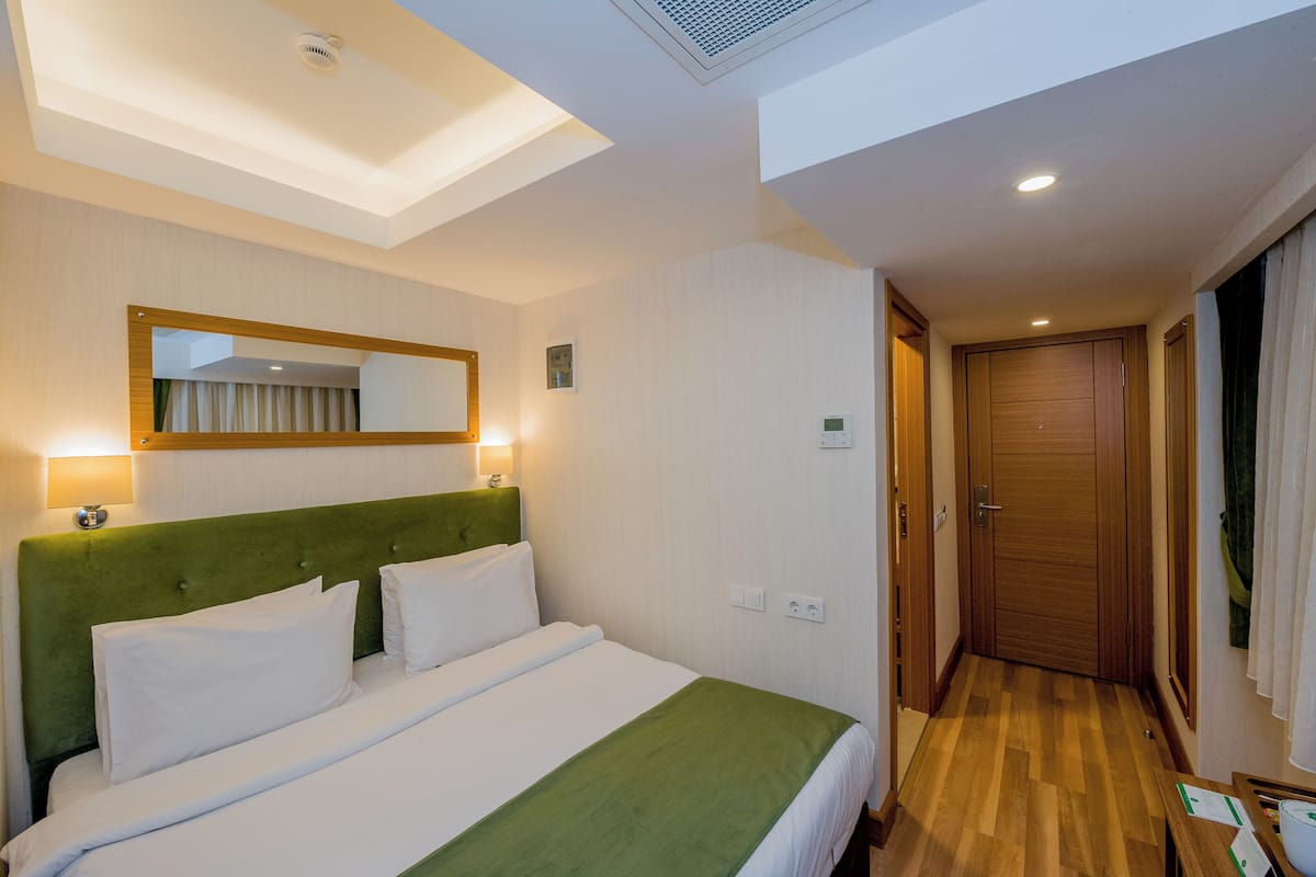 Una habitación de hotel con una cama, cabecera verde, un espejo y una puerta de madera.