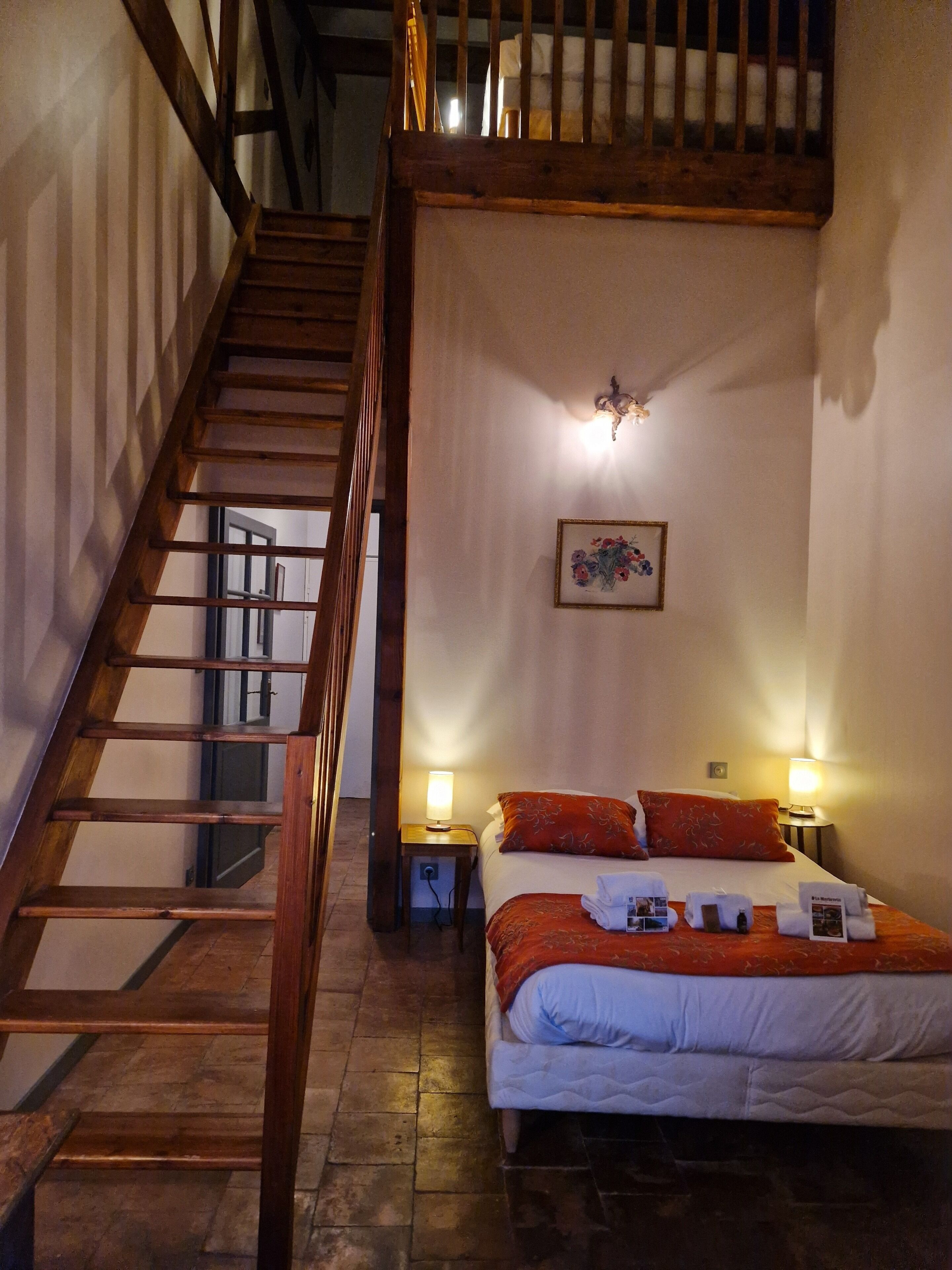Un dormitorio con una escalera de madera, una cama con ropa de cama roja y blanca, dos lámparas de noche y un cuadro en la pared.
