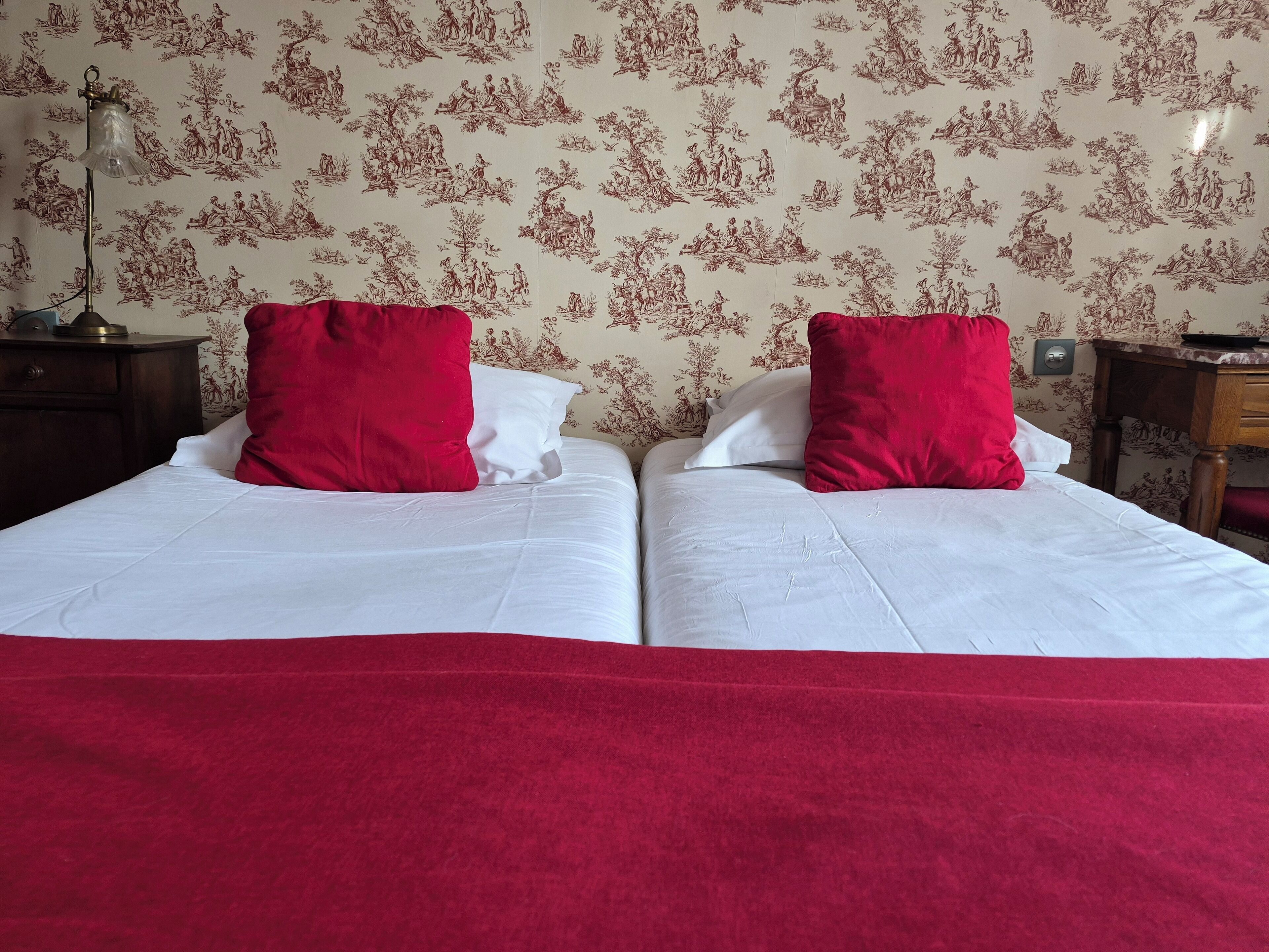 A double bed with white linens and two red pillows, a nightstand with a lamp, and a wall with floral wallpaper.