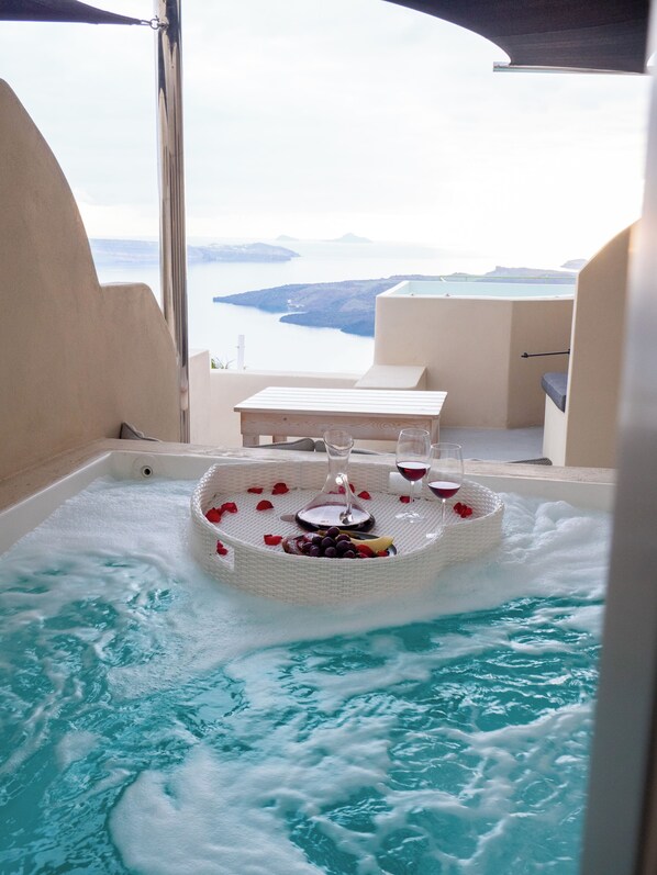 A hot tub with rose petals and wine glasses, surrounded by a view of the sea and hills. - Caldera Cliff Illusion Suites (Santorini)