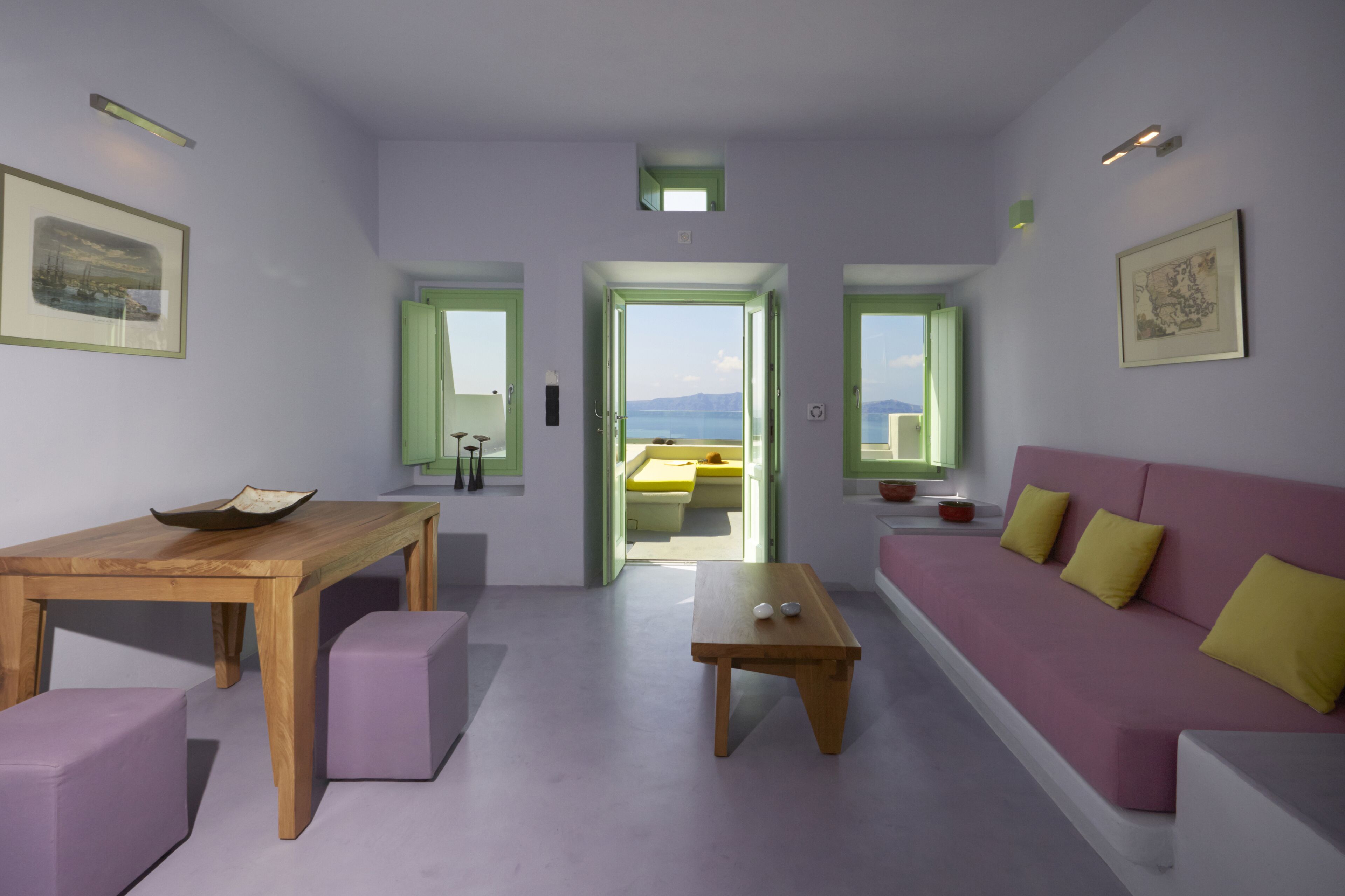 A modern living room with a wooden table, purple ottomans, a pink sofa with yellow cushions, and a view of the sea through a doorway.