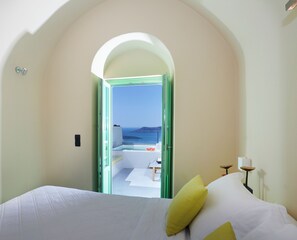 A bedroom with a bed, a view of the sea through an open door, and a small table with a lamp. - Caldera Cliff Illusion Suites (Santorini)