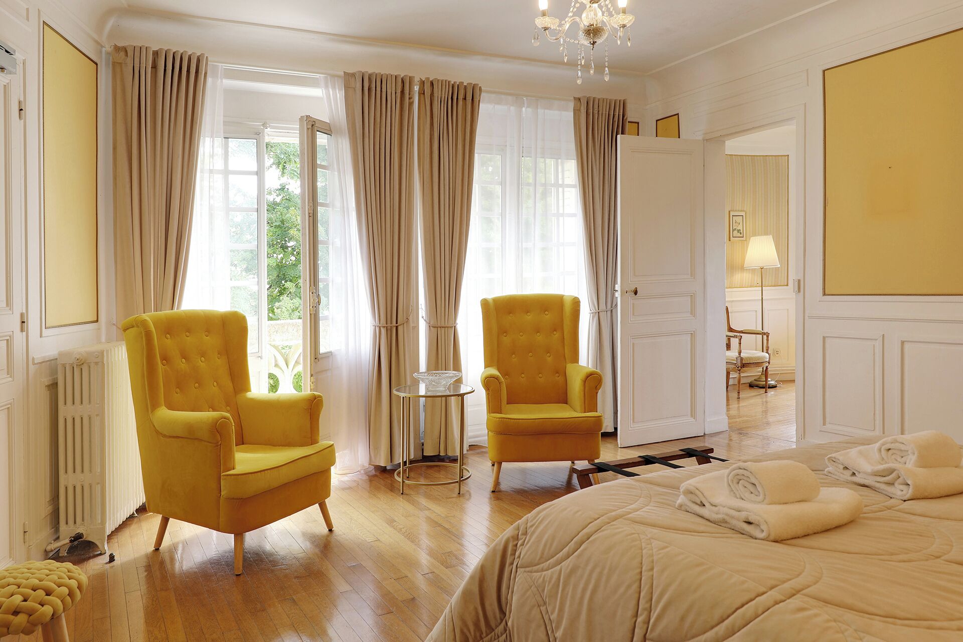 A room with two yellow armchairs, a bed with beige bedding, a chandelier, and a view through large windows with curtains.