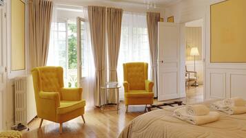 A room with two yellow armchairs, a bed with beige bedding, a chandelier, and a view through large windows with curtains.