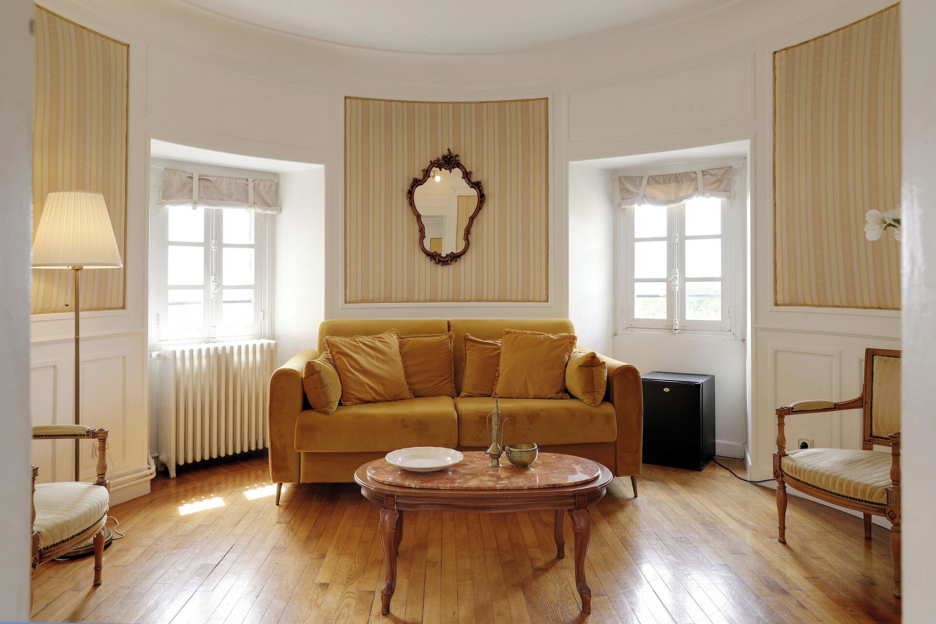 Ein Zimmer mit einer Couch, einem runden Tisch, einem Sessel, einer Heizung und einem großen Spiegel an der Wand.