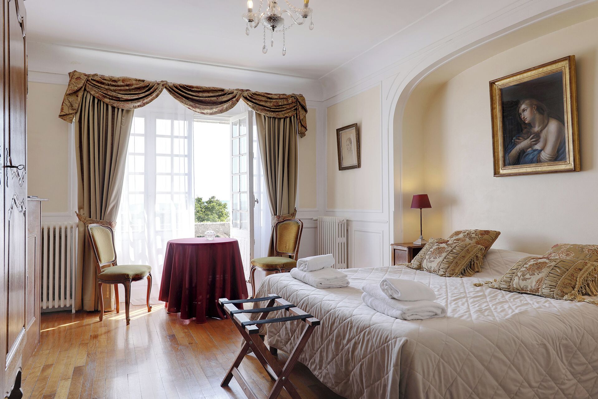 A bedroom with a bed, two chairs, a table, a radiator, and a painting on the wall.