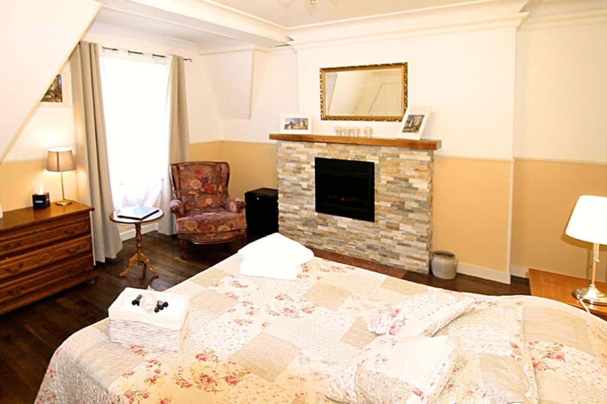 A hotel room with a bed, a chair, a dresser, a lamp, a fireplace, and a mirror on the wall.
