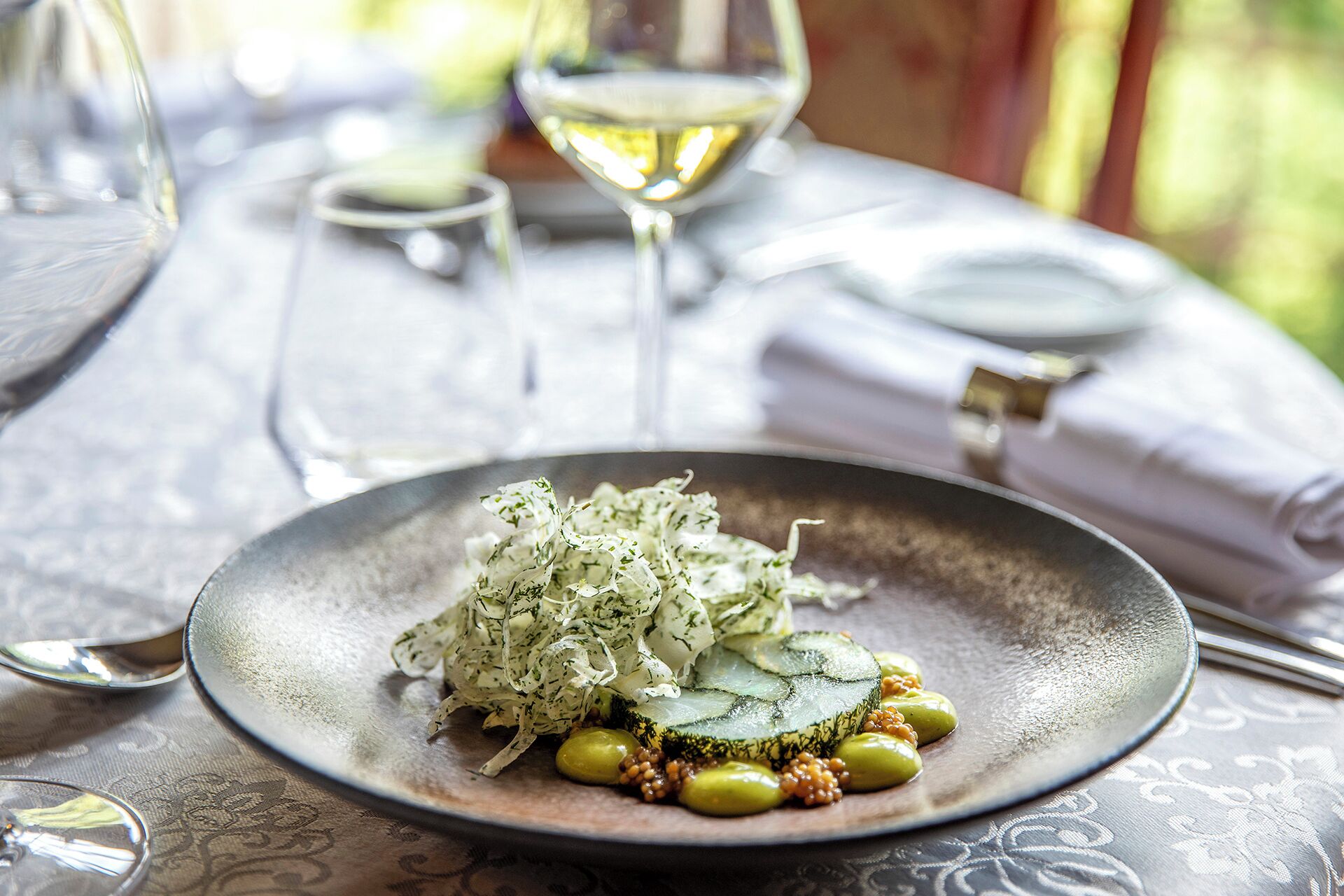 Un plato de comida con pepinos en rodajas, salsa verde y aceitunas sobre una mesa con una copa de vino y una servilleta doblada.