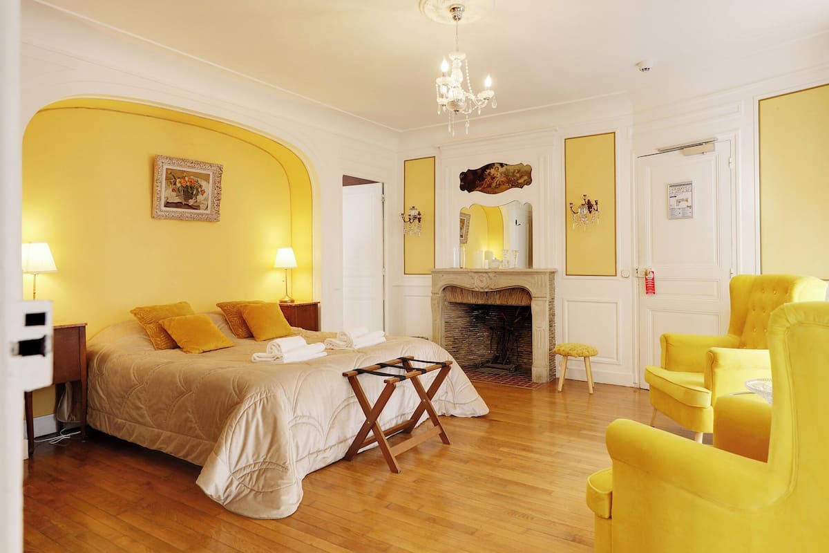 A hotel room with a bed, a fireplace, a chandelier, and yellow chairs.