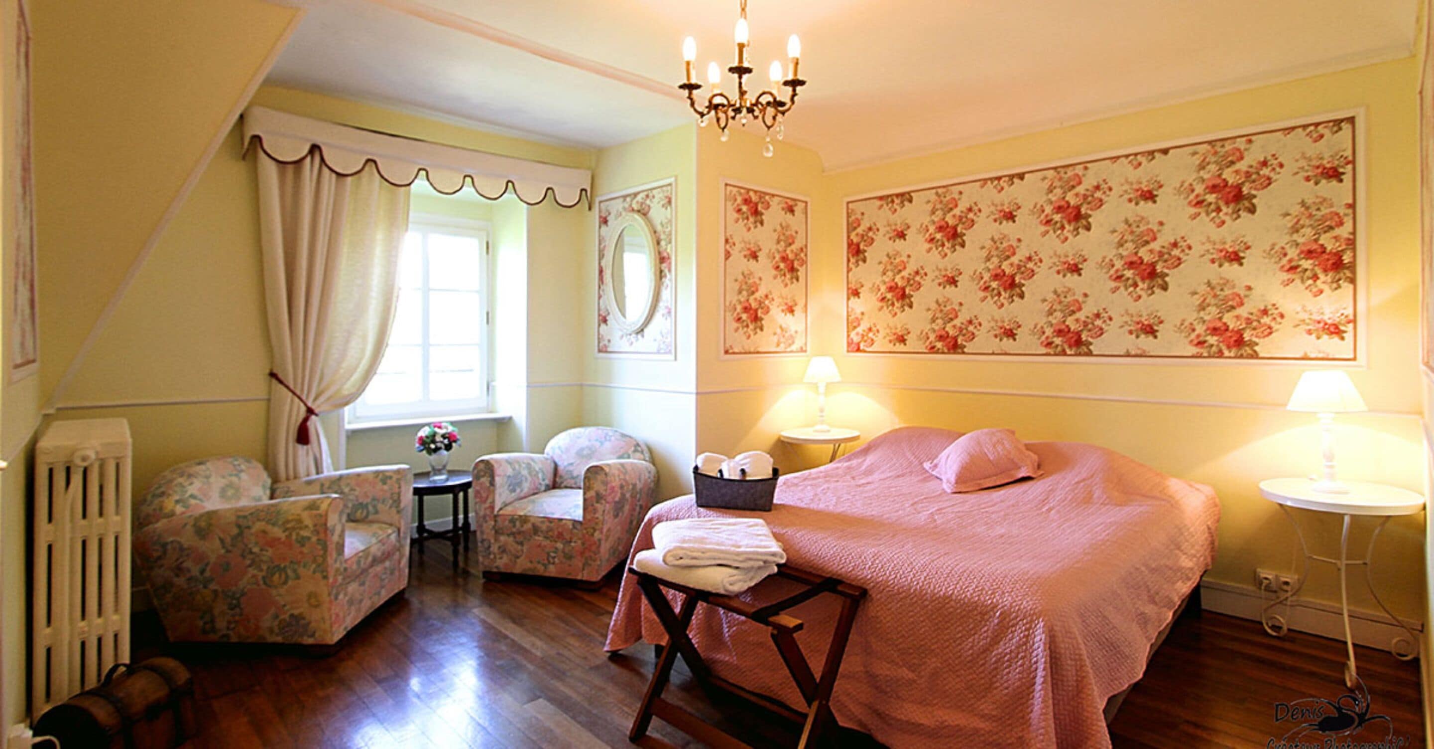 Une chambre à coucher avec un lit, deux chaises, une table avec un panier, un radiateur et un papier peint à motifs floraux.