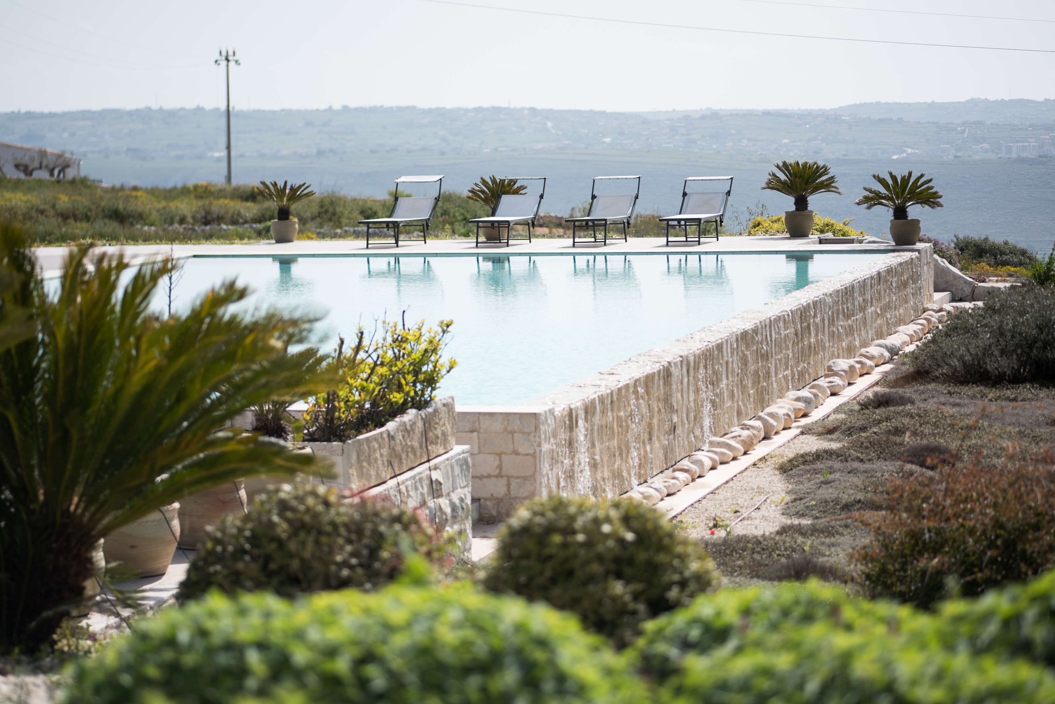 Une piscine entourée de verdure, agrémentée de chaises longues et de plantes en pot, et offrant une vue sur une étendue d’eau.