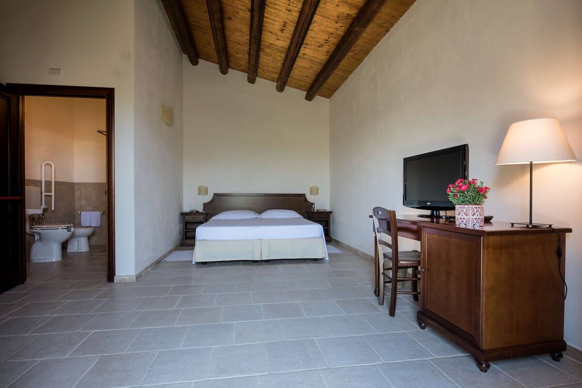 A bedroom with a bed, a desk with a computer, a TV, and a lamp.