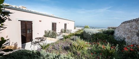 Un edificio con un muro de piedra, un patio con mesa y sillas, y un jardín con diversas plantas y flores.