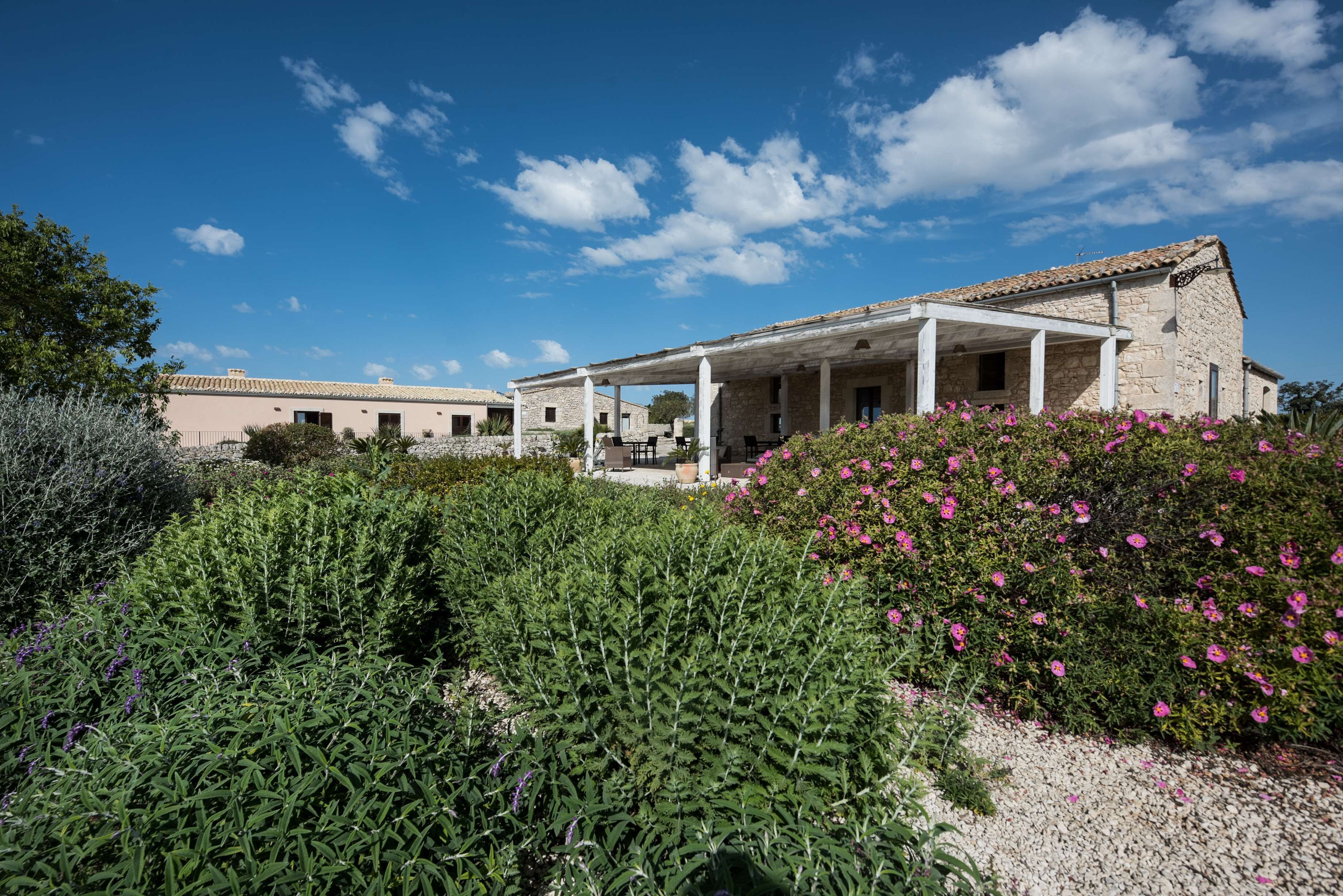 A stone building with a covered patio and a garden with green shrubs and pink flowers.