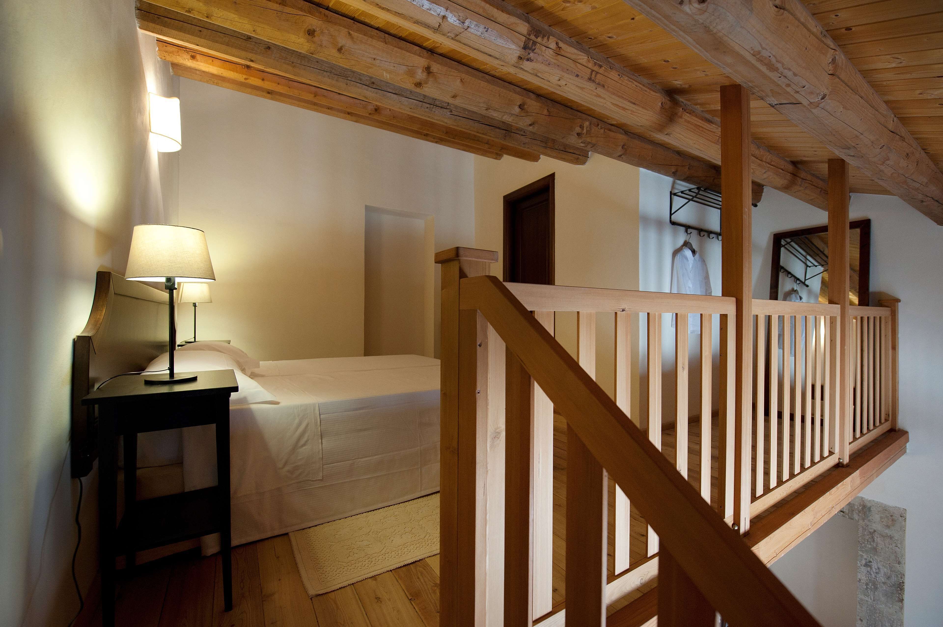 A bedroom with a wooden ceiling, a bed, a nightstand with a lamp, and a wooden railing.