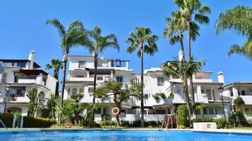 A pool area with a clear blue pool, surrounded by palm trees and white buildings with balconies.