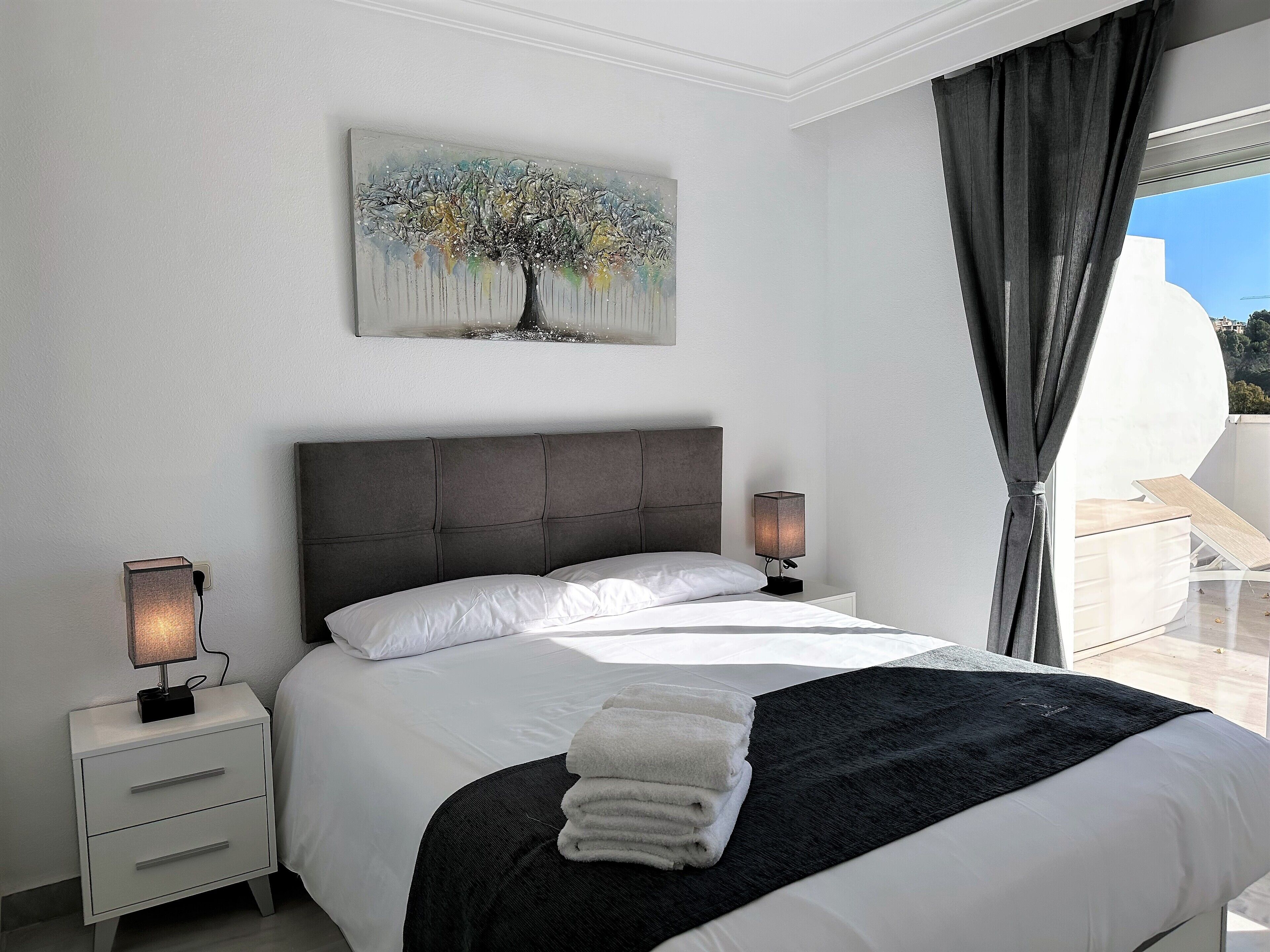 A bedroom with a large bed, two bedside tables, a painting, and a view of the outdoors.
