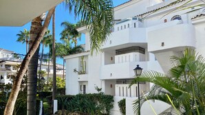 Un edificio blanco con balcones, un jardín con fuente de agua y palmeras.