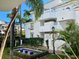 A white building with balconies, a garden with a water feature, and palm trees.