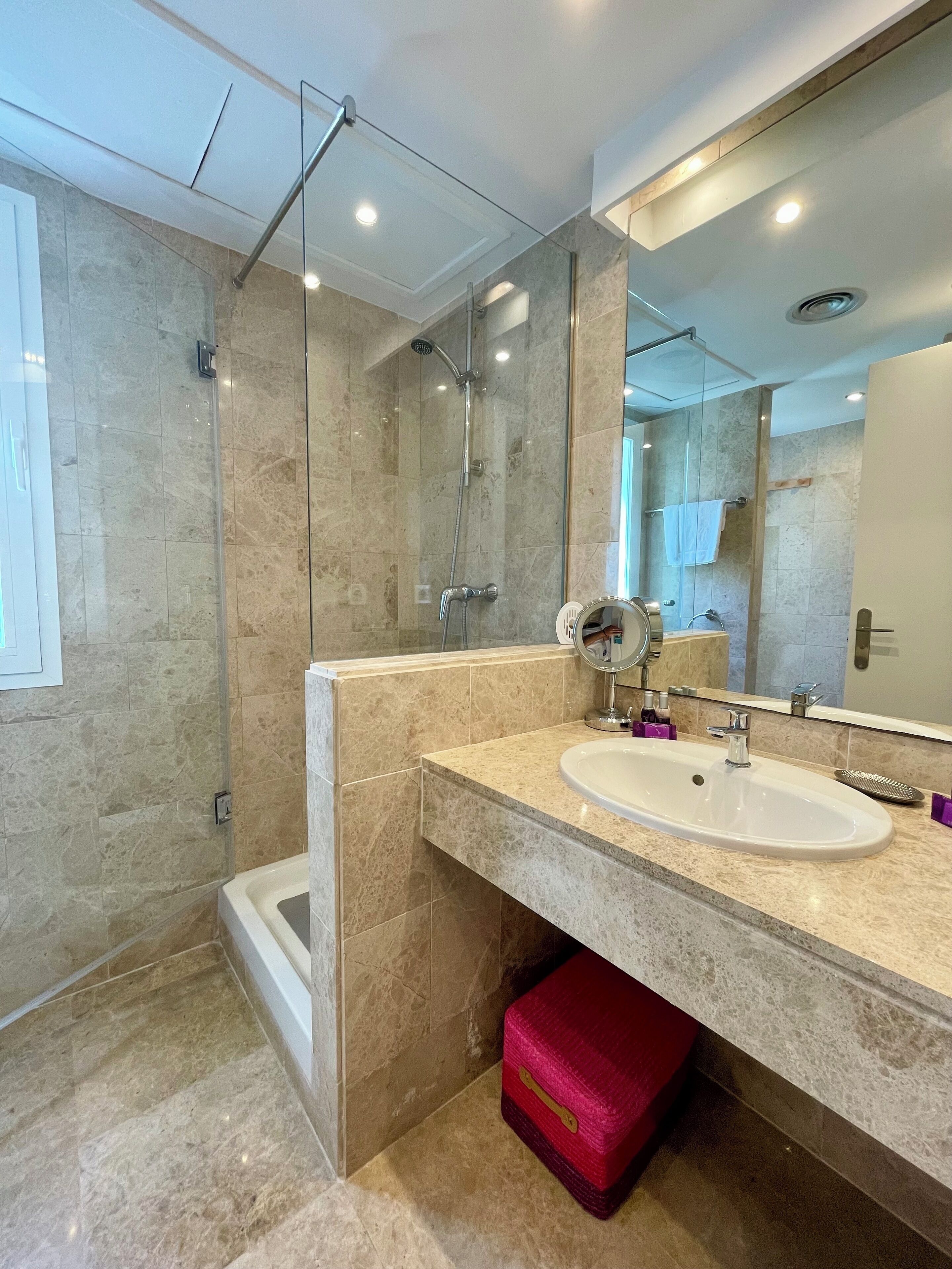 A modern bathroom with a glass-enclosed shower, a large mirror, and a marble-tiled floor.