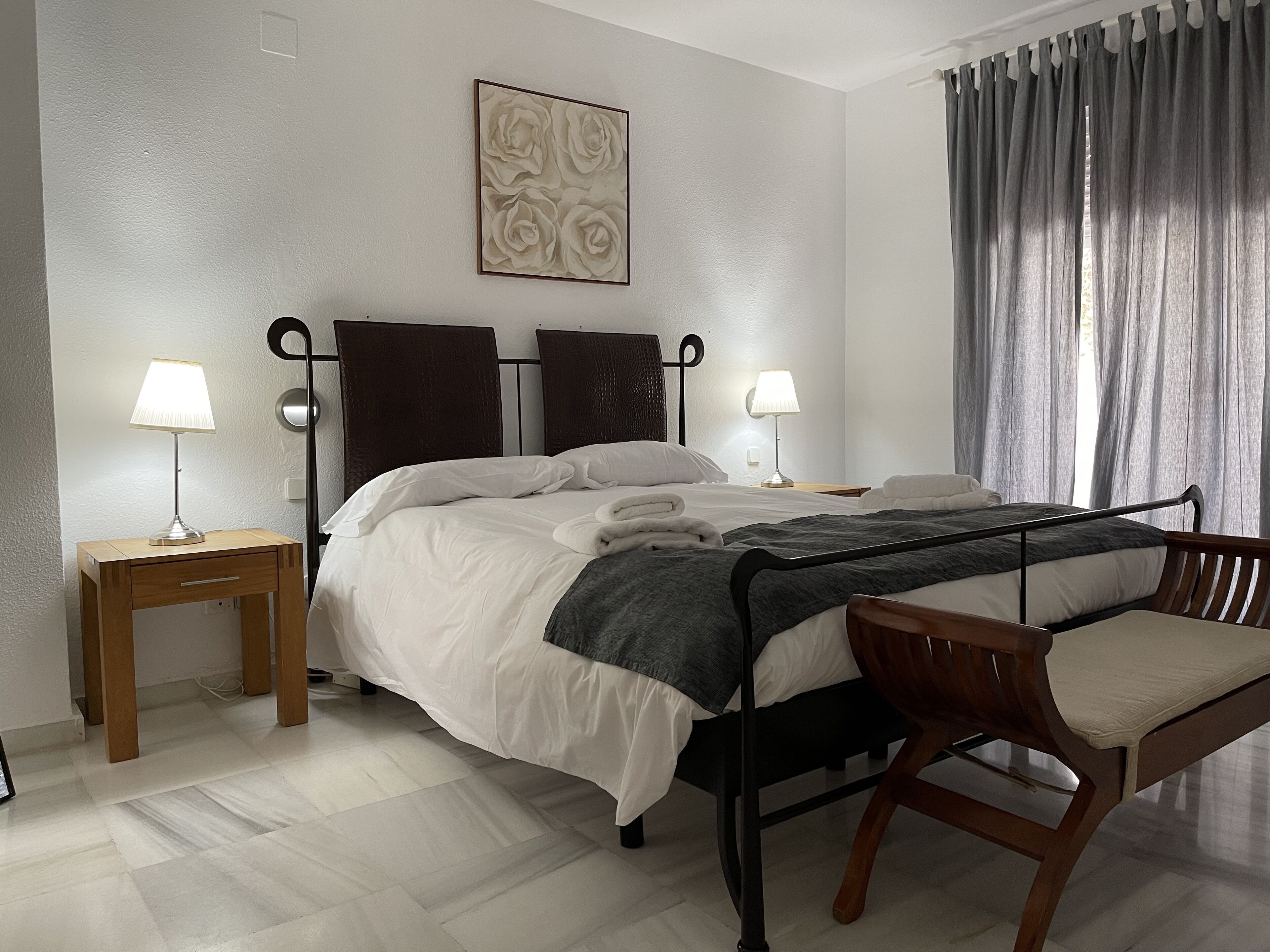 A neatly arranged hotel room with a bed, two bedside tables with lamps, a chair, a painting on the wall, and a window with curtains.