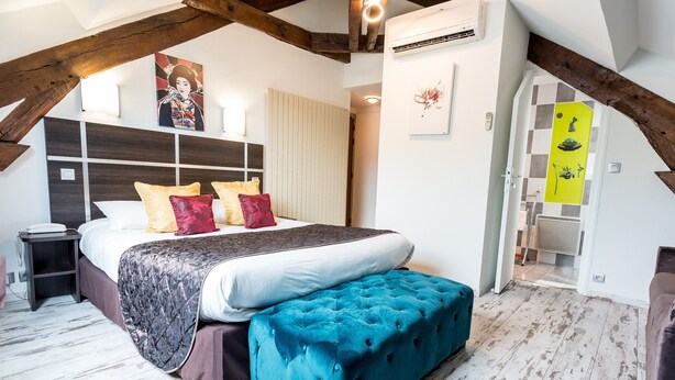 A bedroom with a large bed, a tufted ottoman, wooden beams, a headboard, and a wall-mounted air conditioner. - Hotel du Centre (Gramat)