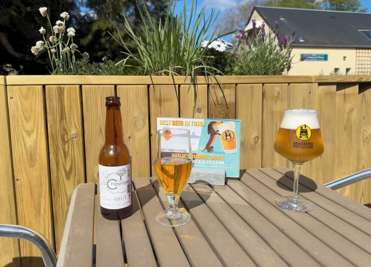 Une table en bois avec une bouteille et deux verres de bière, un panneau indicateur et une clôture en bois ornée de plantes.