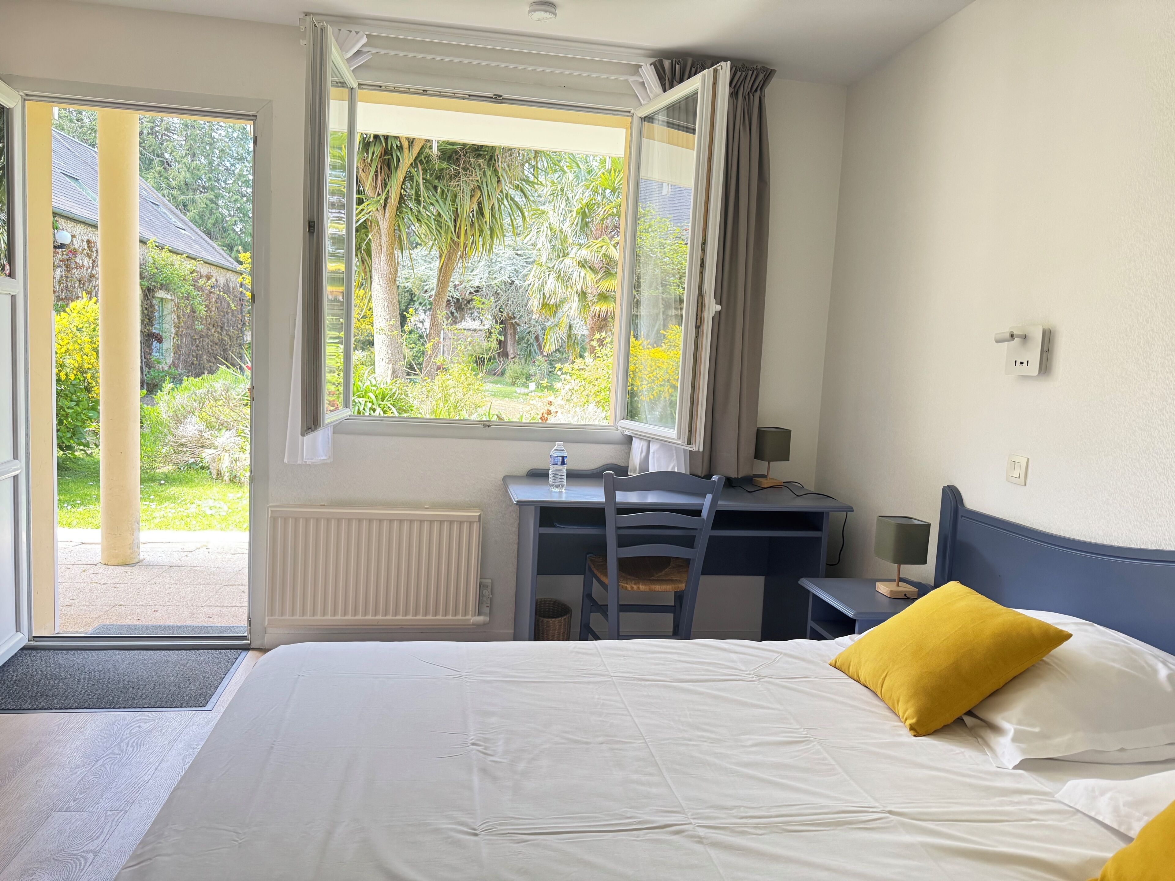 Une chambre à coucher comprenant un lit, un bureau, une chaise, une fenêtre avec des rideaux et une vue sur la verdure à l’extérieur.